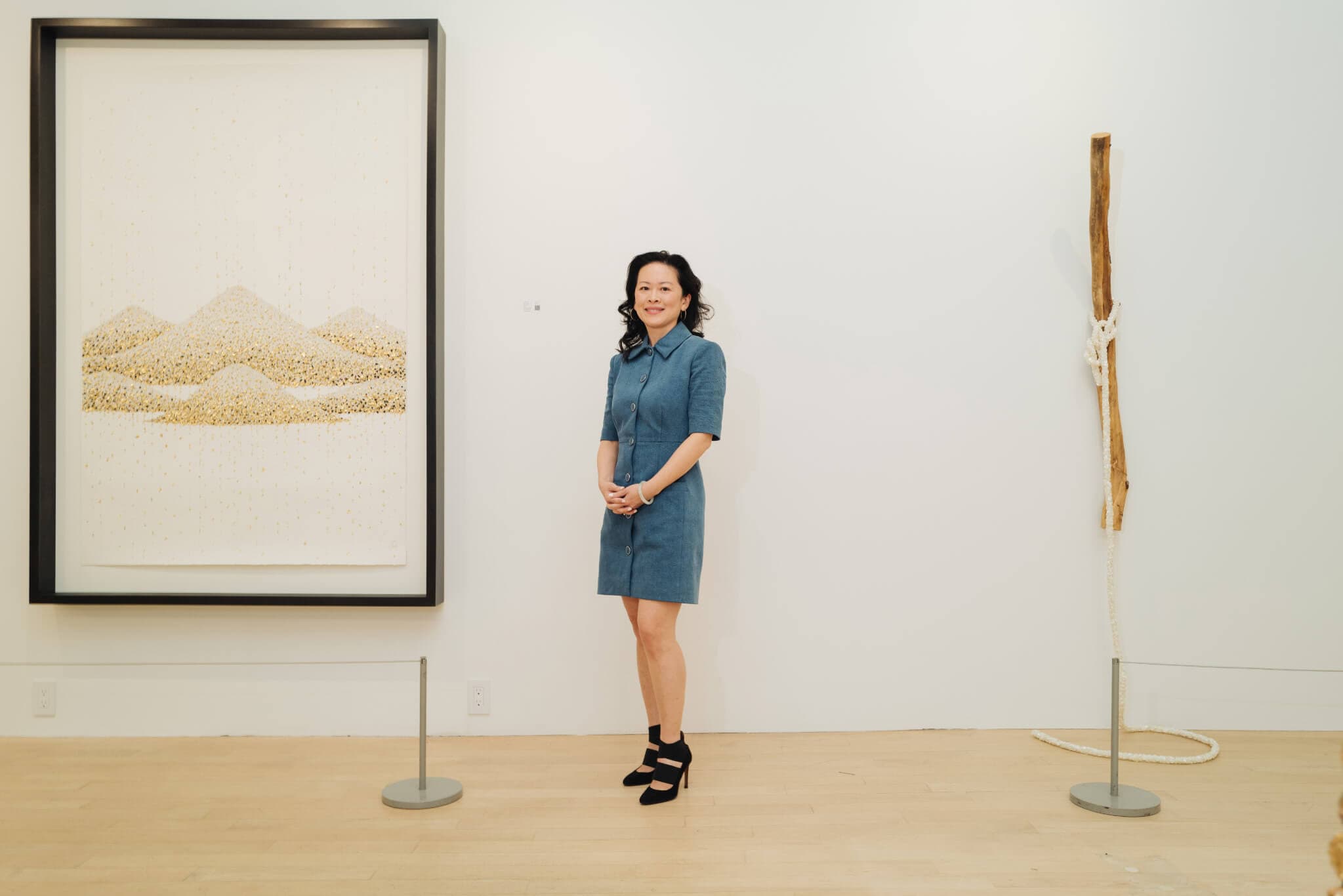 A woman wearing a blue dress stands beside a hanging, framed image of piles of stones.