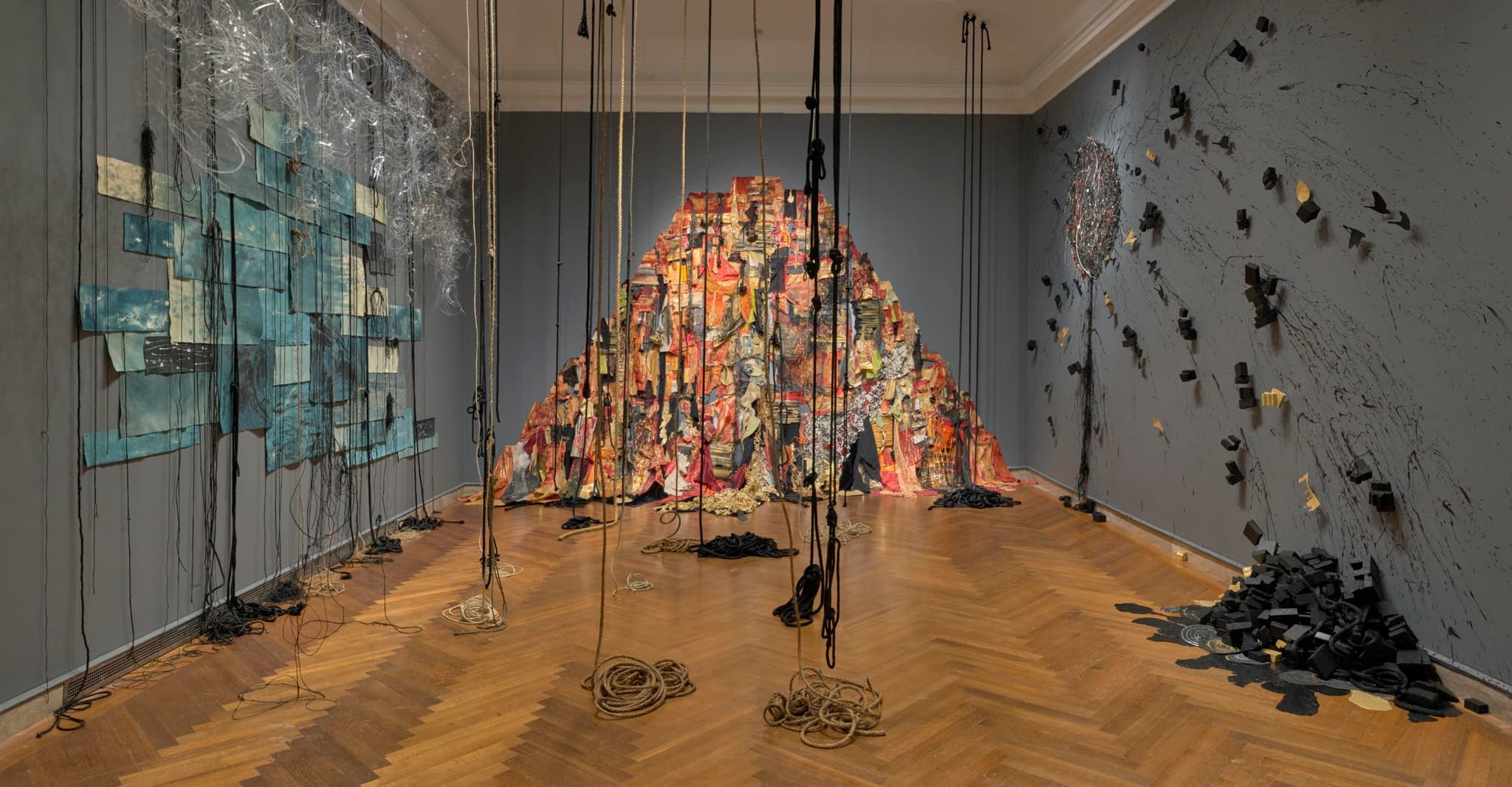 Heidi Whitman's installation features ropes dangling from the ceiling, while red, orange, and black hued art takes form on one side of the wall.