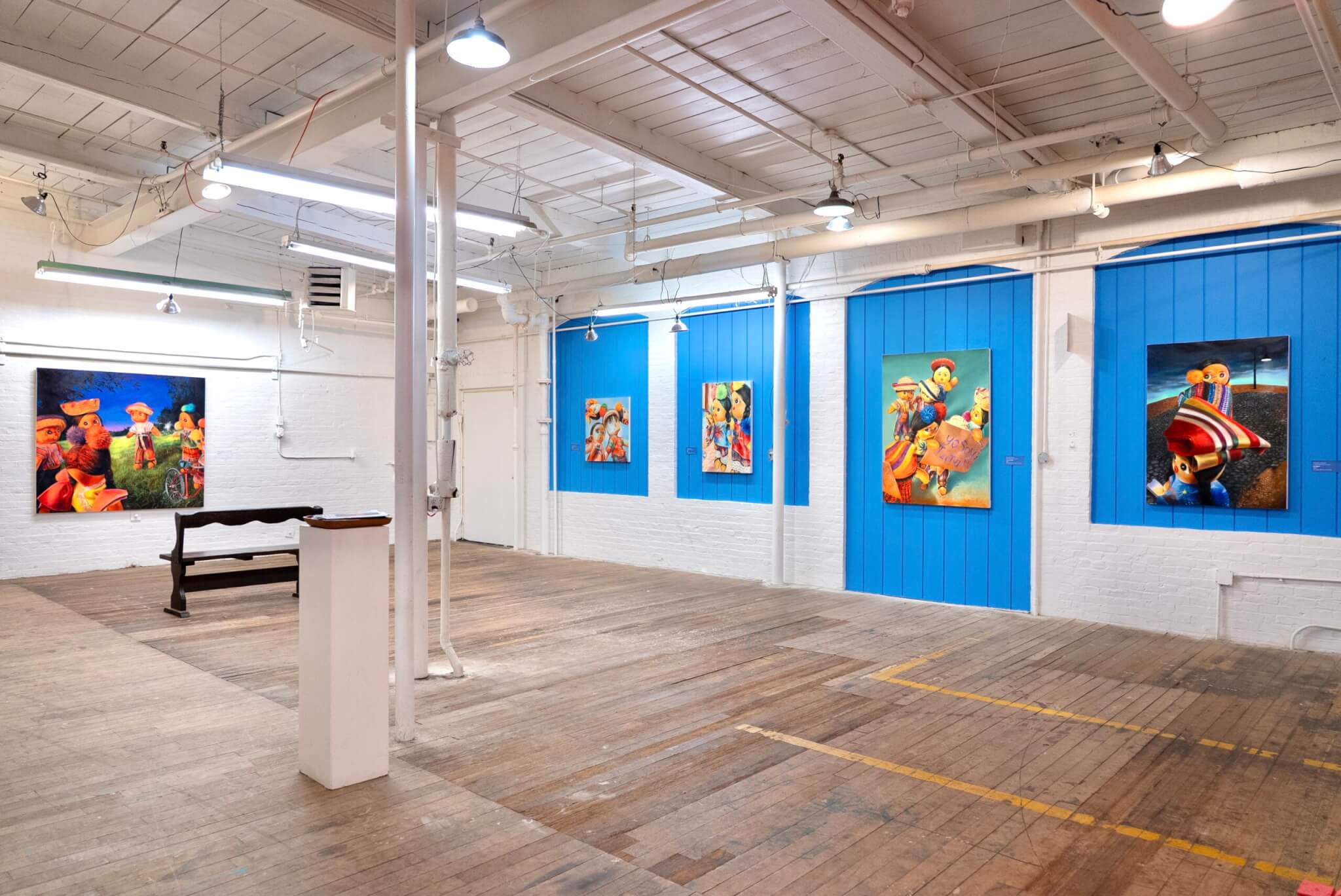 Vibrant paintings of worry dolls hang on the blue walls of a gallery space.